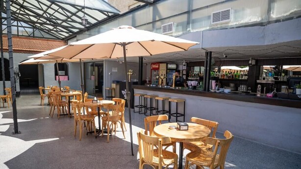 An outdoor café area with wooden tables and chairs, umbrellas, and a bar with various bottles. - Dali House (Amman)