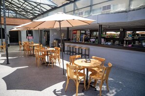 Een terras met houten tafels en stoelen, parasols en een bar met diverse flessen.