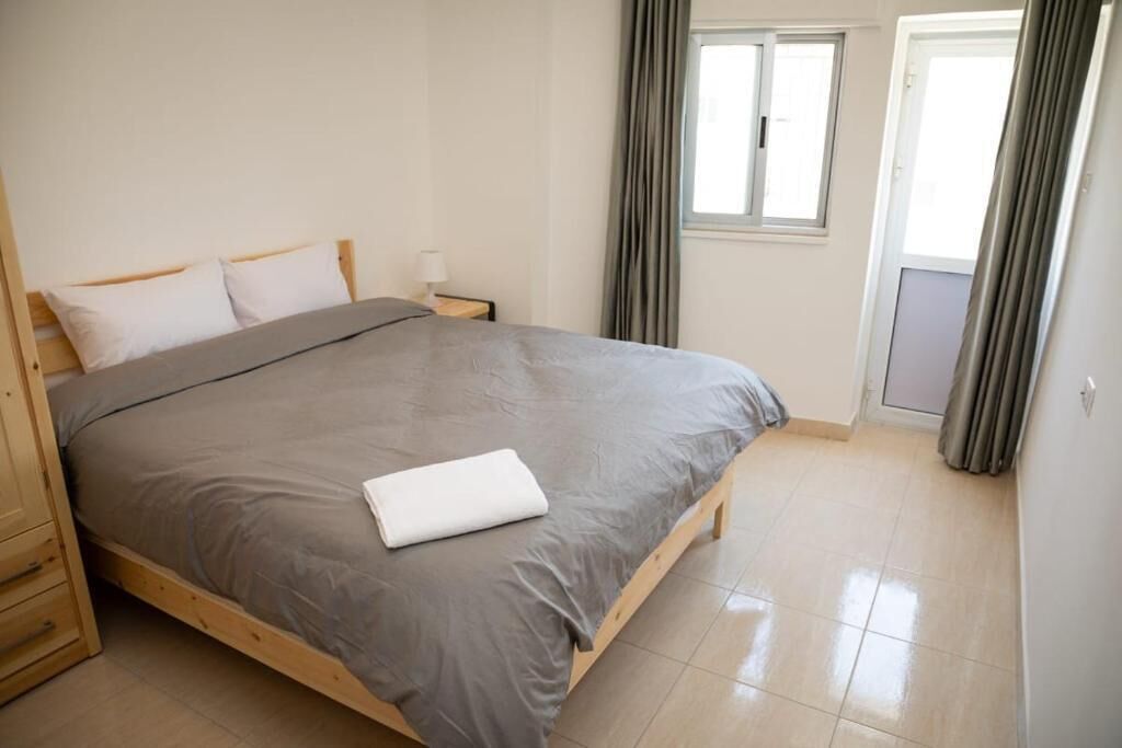 A neatly made bed with a grey bedspread and white pillows, a wooden nightstand with a lamp, and a window with curtains.