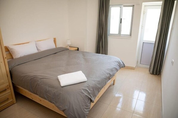 A neatly made bed with a grey bedspread and white pillows, a wooden nightstand with a lamp, and a window with curtains.