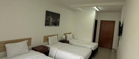 A hotel room with three beds, a nightstand with a lamp, a painting on the wall, and a wooden door.