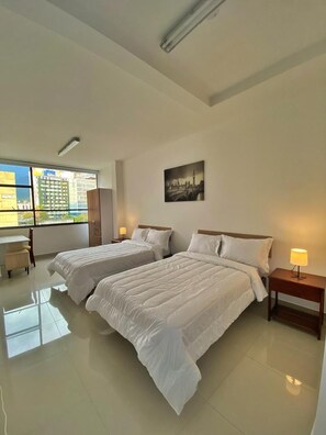 A hotel room with two beds, a desk, a chair, a window with a cityscape view, and a painting on the wall. - HOSPEDAJE BOGOTA CENTRO  (Bogotá)