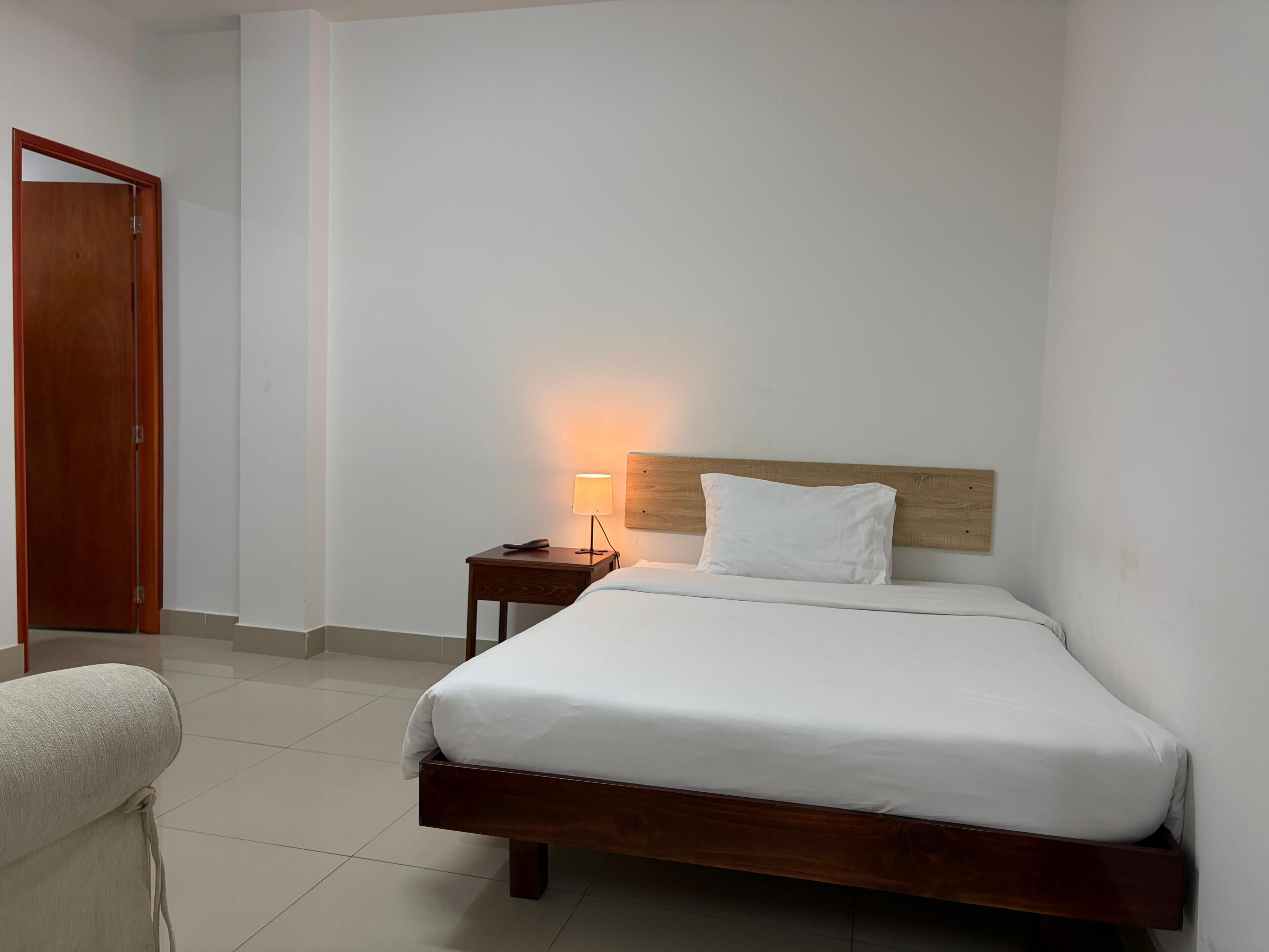 A hotel room with a wooden bed, a bedside table with a lamp, and a door.