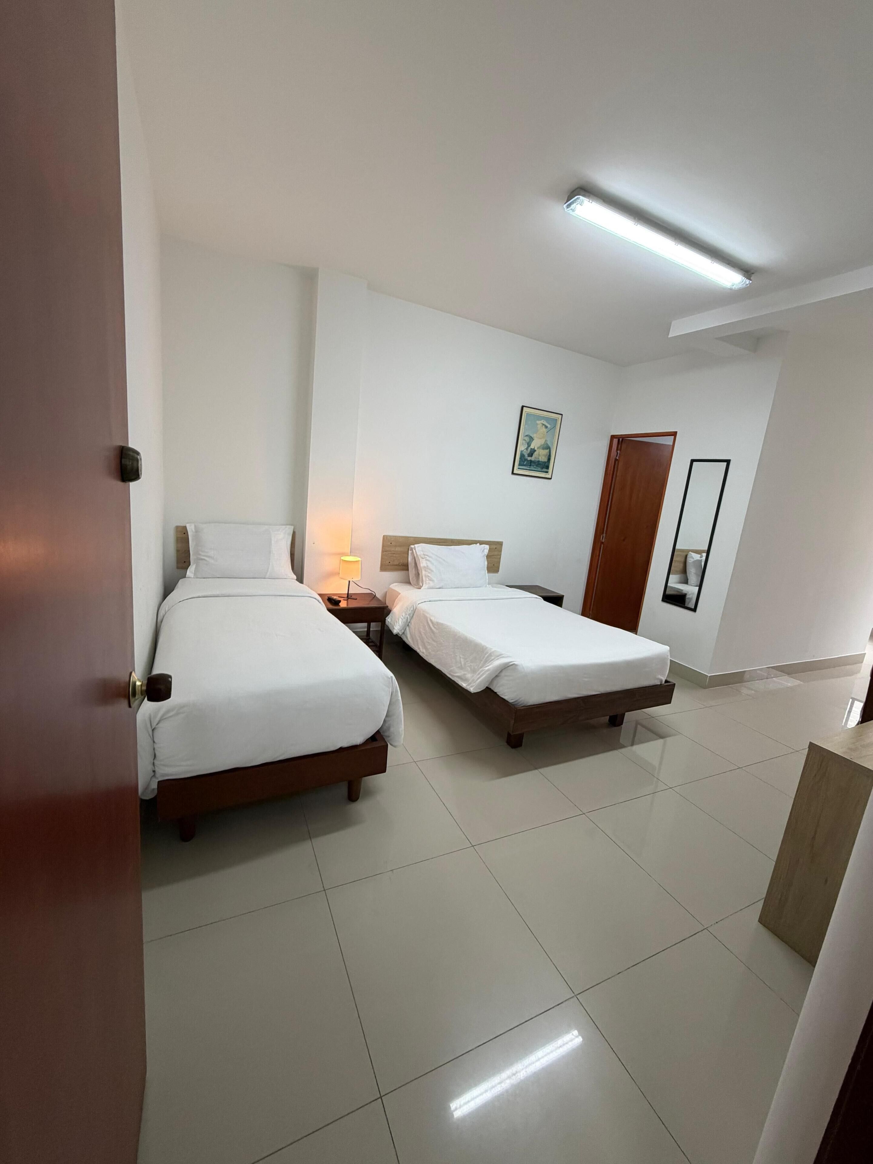 A hotel room with two beds, a nightstand, a wall-mounted lamp, a framed picture, a mirror, and a door.