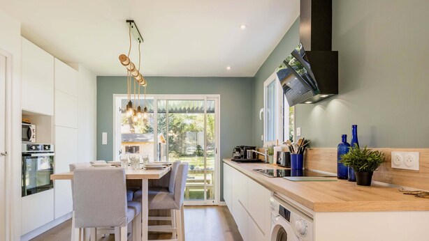 A modern kitchen with a dining area, built-in appliances, and a large window offering a view of the outdoors. - Charming Renovated House Near Beach with Garden & Parking (Saint-Brevin-les-Pins)