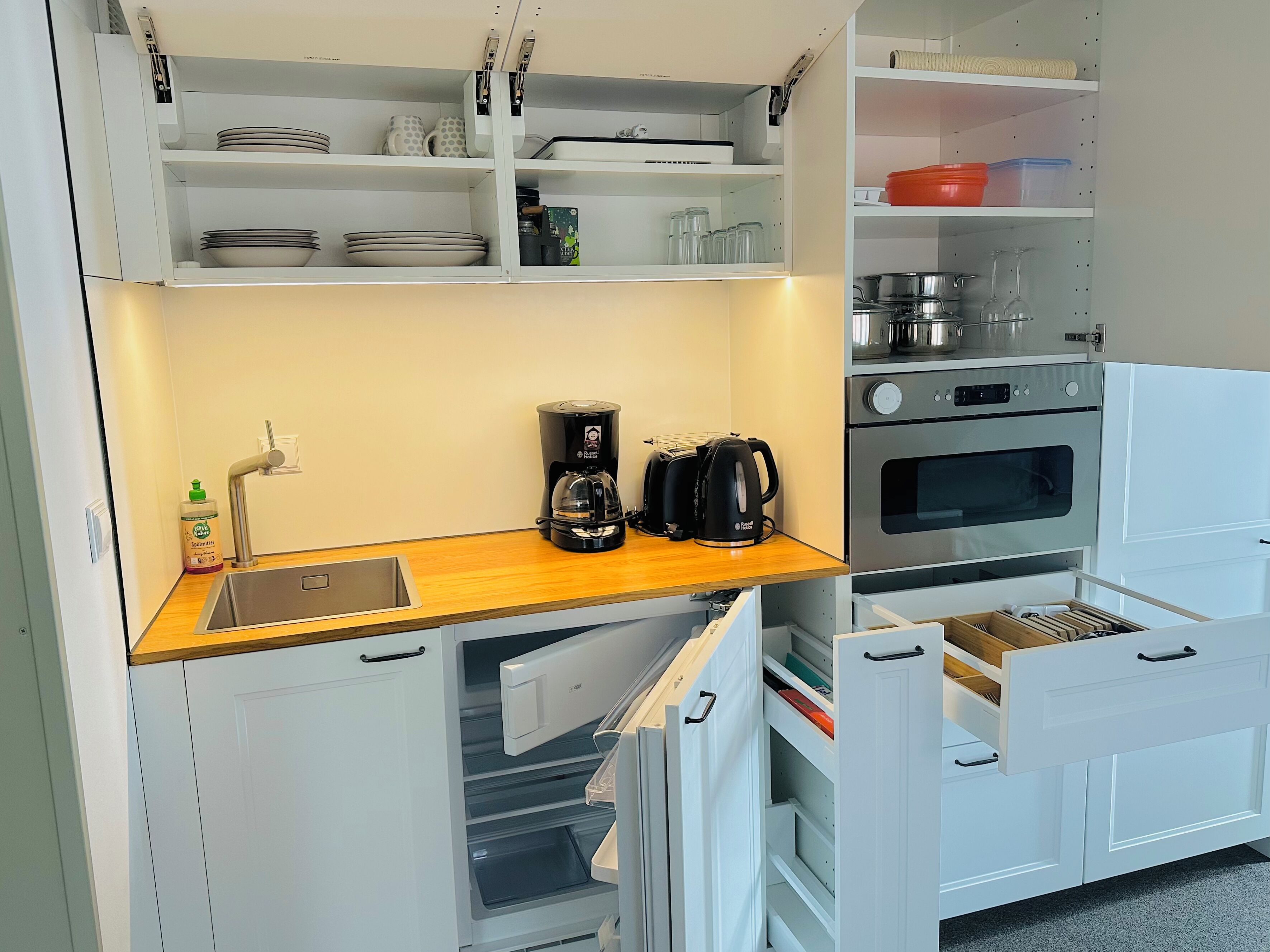Una cocina compacta con armarios blancos, encimera de madera, cafetera, hervidor y lavabo.