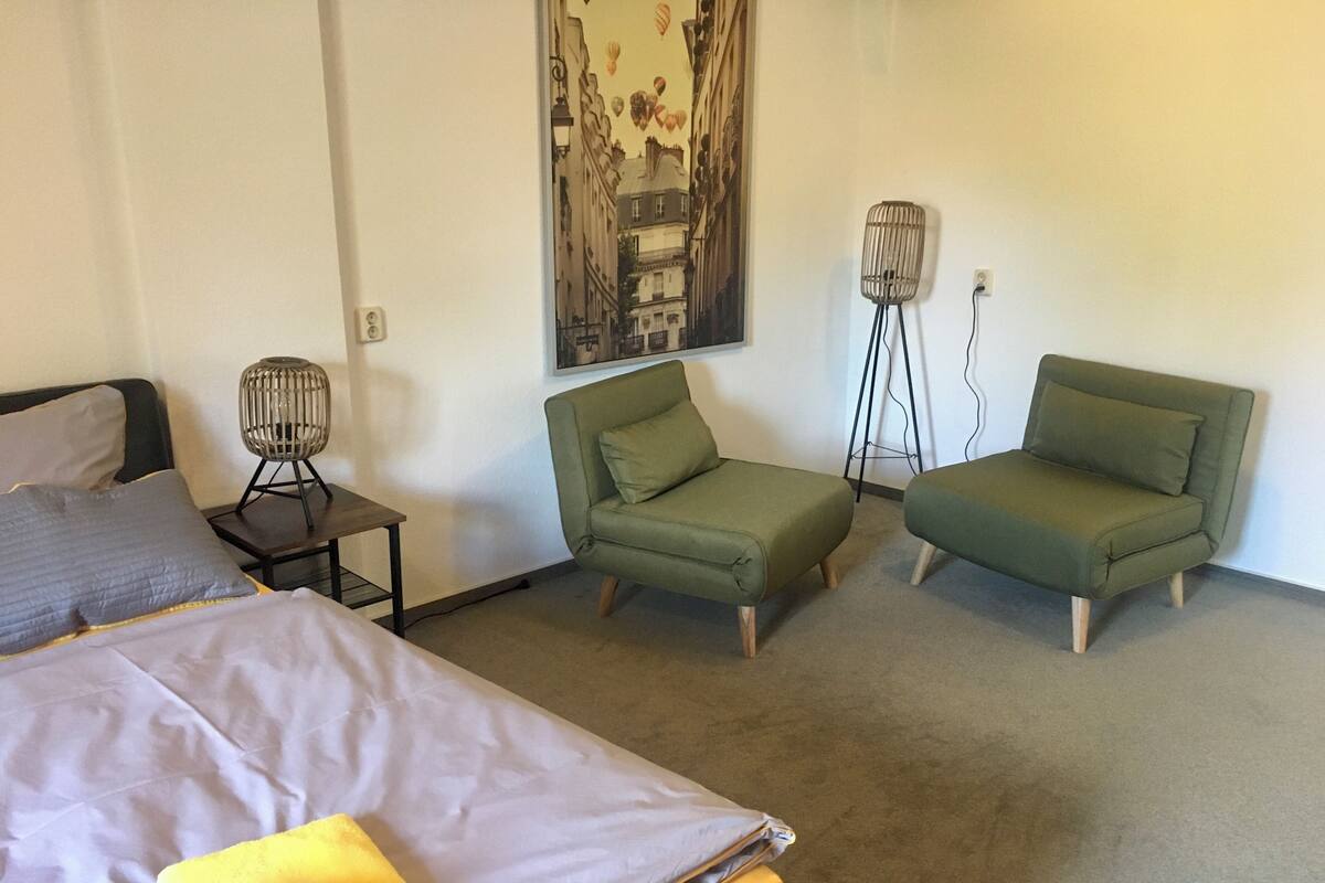 Una habitación con una cama, dos sillones verdes, una mesita con una lámpara y un cuadro colgado en la pared.