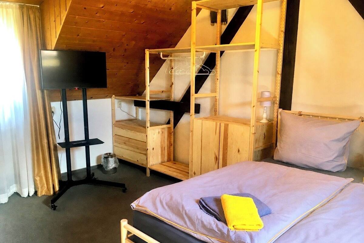 Una cama con dosel de madera, un televisor sobre un mueble bajo, un escritorio y una silla en una habitación con techo inclinado.