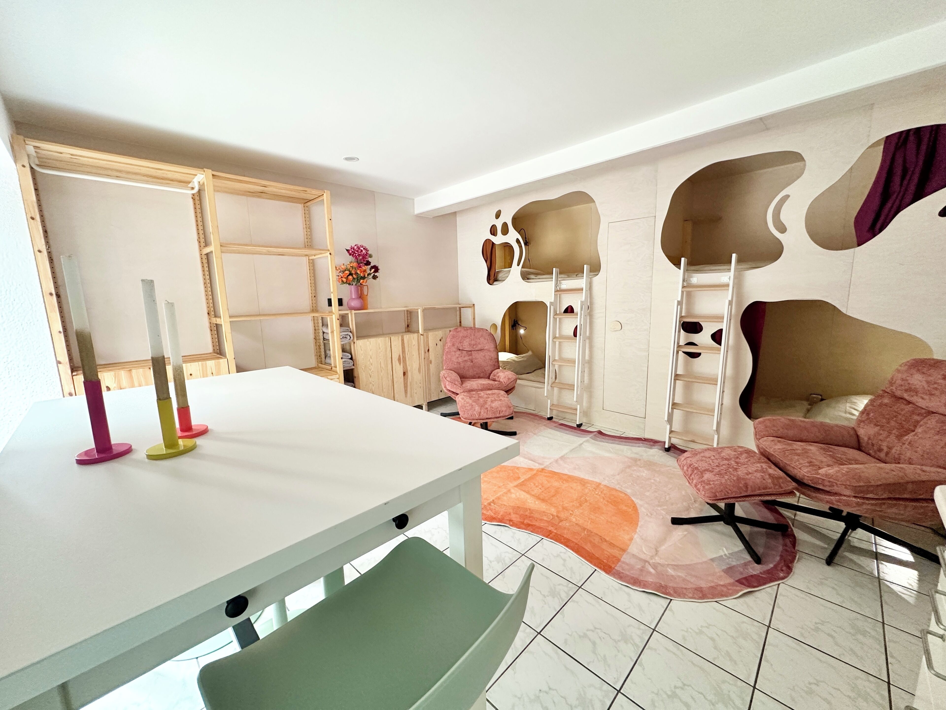 A room with a white table, pink chairs, a green chair, a white shelf, a pink vase with flowers, a ladder, and a unique wall design.