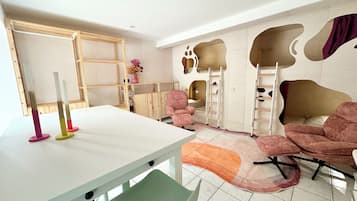 A room with a white table, pink chairs, a green chair, a white shelf, a pink vase with flowers, a ladder, and a unique wall design.