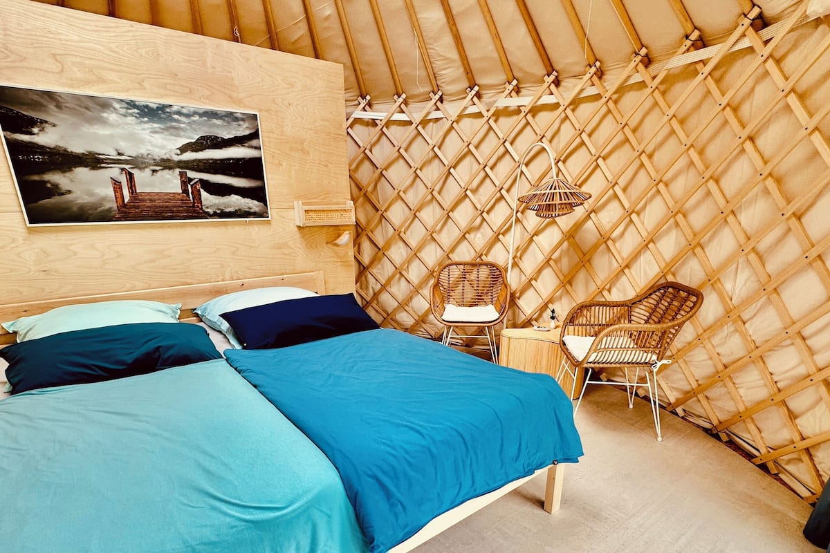 Una habitación con una cama, dos sillas, una mesita pequeña, un cuadro colgado en la pared y un muro de celosía.