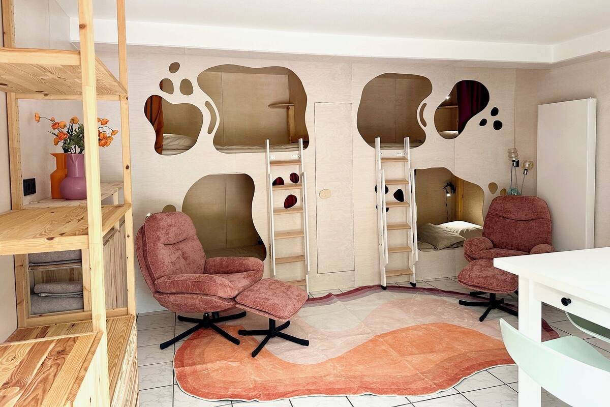 Una habitación con una cama litera, dos sillones rosa, un escritorio blanco y una estantería de madera.