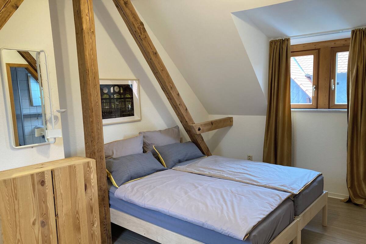 Un dormitorio con techo inclinado, vigas de madera, cama con almohadas, mesita de noche y ventana con cortinas.