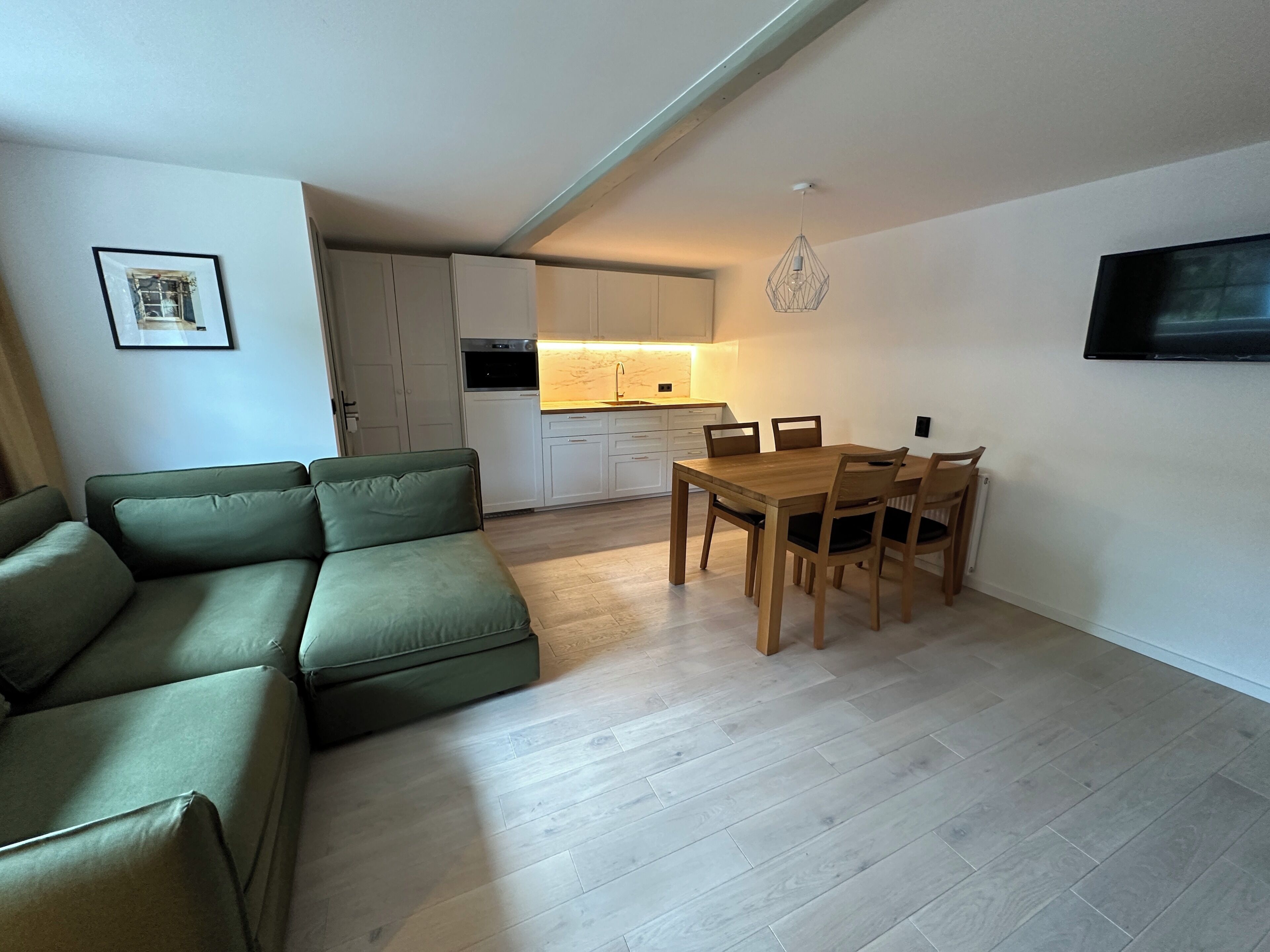 A modern living space with a sectional sofa, a dining table with chairs, a kitchen area, and a mounted TV.