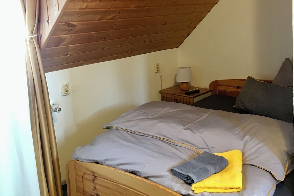 Un dormitorio con techo inclinado, una cama con colchón gris y azul, cabecero de madera, mesita de noche con lámpara y una ventana con cortinas.