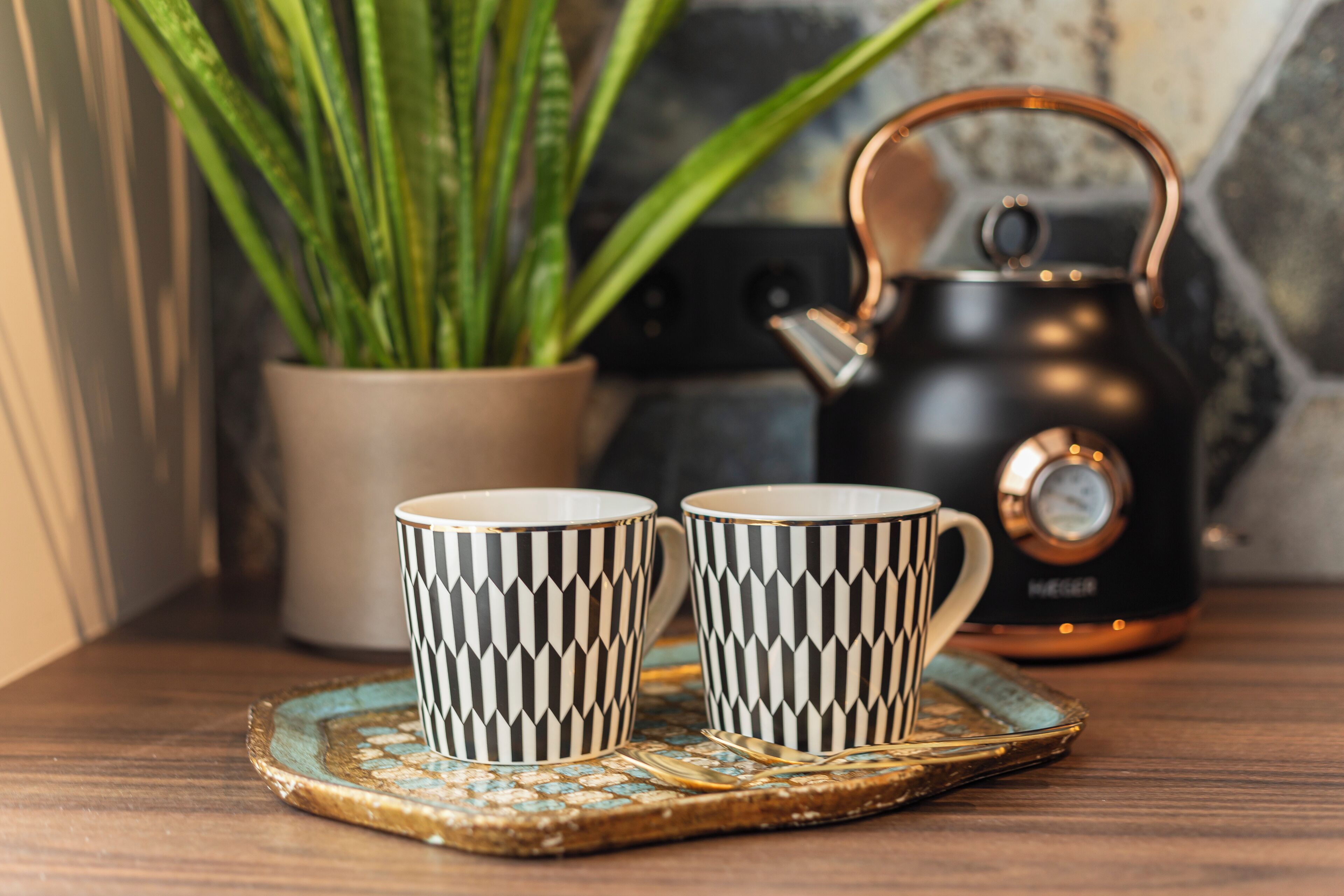 Twee versierde kopjes op een onderzetter, een bloempot en een waterkoker op een tafel.