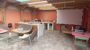 A rustic outdoor dining area with wicker chairs, a wooden table, and a bar area with a large screen.