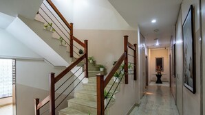 Un escalier avec des rampes en bois et des plantes en pot, menant à un couloir orné de tableaux et surmonté d’une petite table sur laquelle est posé un objet décoratif.