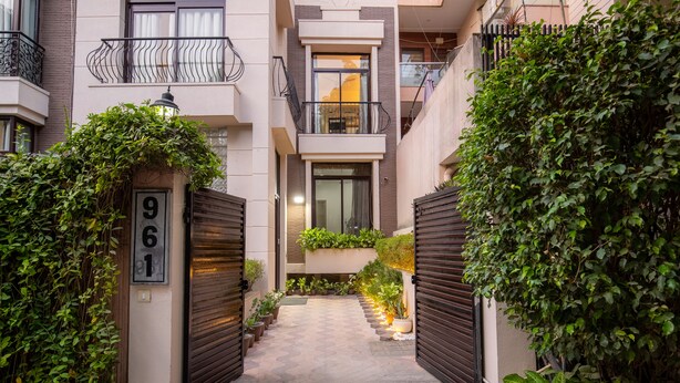 Un étroit sentier mène à un immeuble d’appartements moderne doté d’un balcon, de plantes en pot et d’un portail portant le numéro 961.