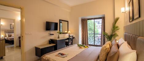 A hotel room with a bed, desk, and TV. There is a window with curtains and a plant on the bedside table.
