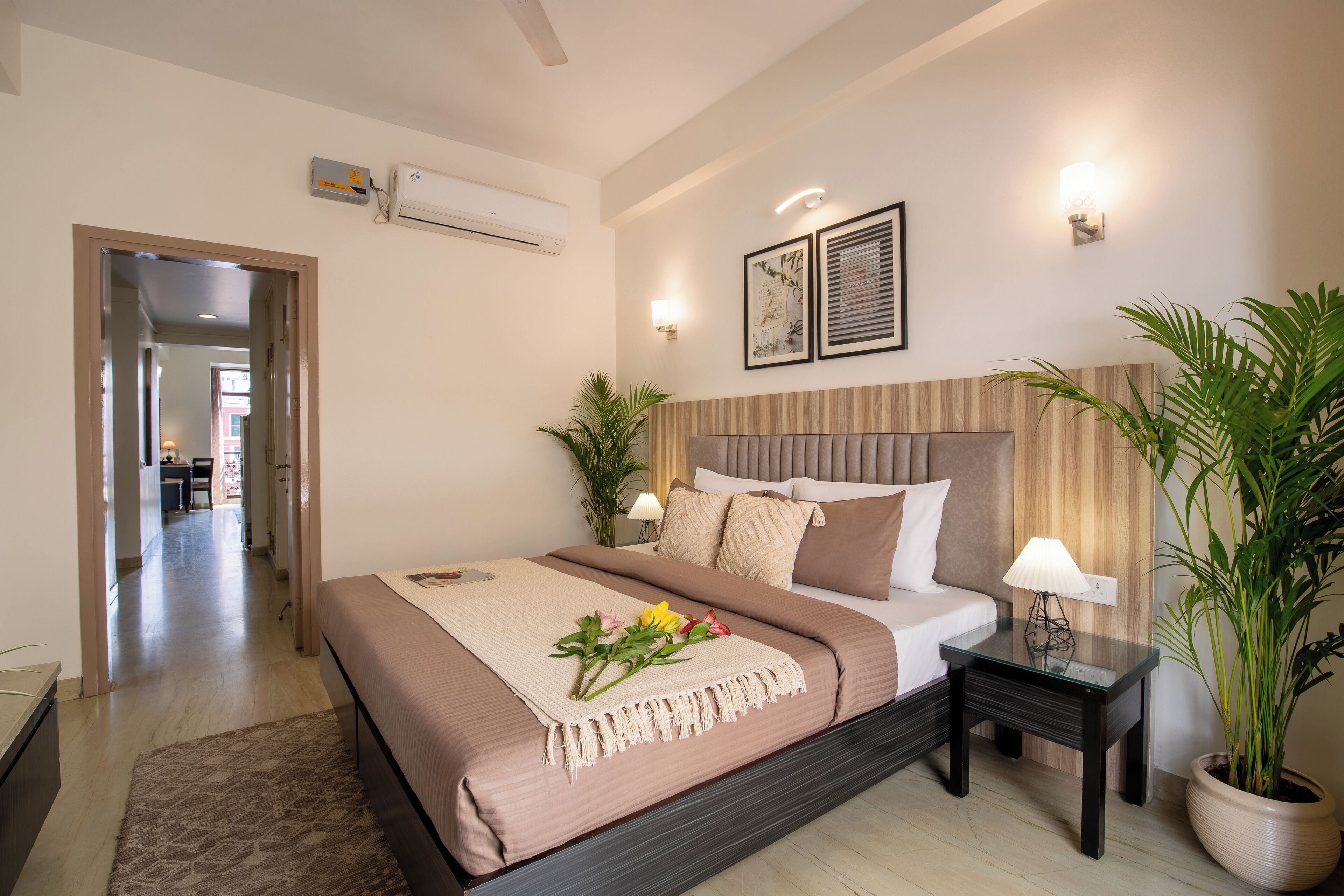 A modern bedroom with a large bed, bedside lamps, a potted plant, and a view into the adjacent room.