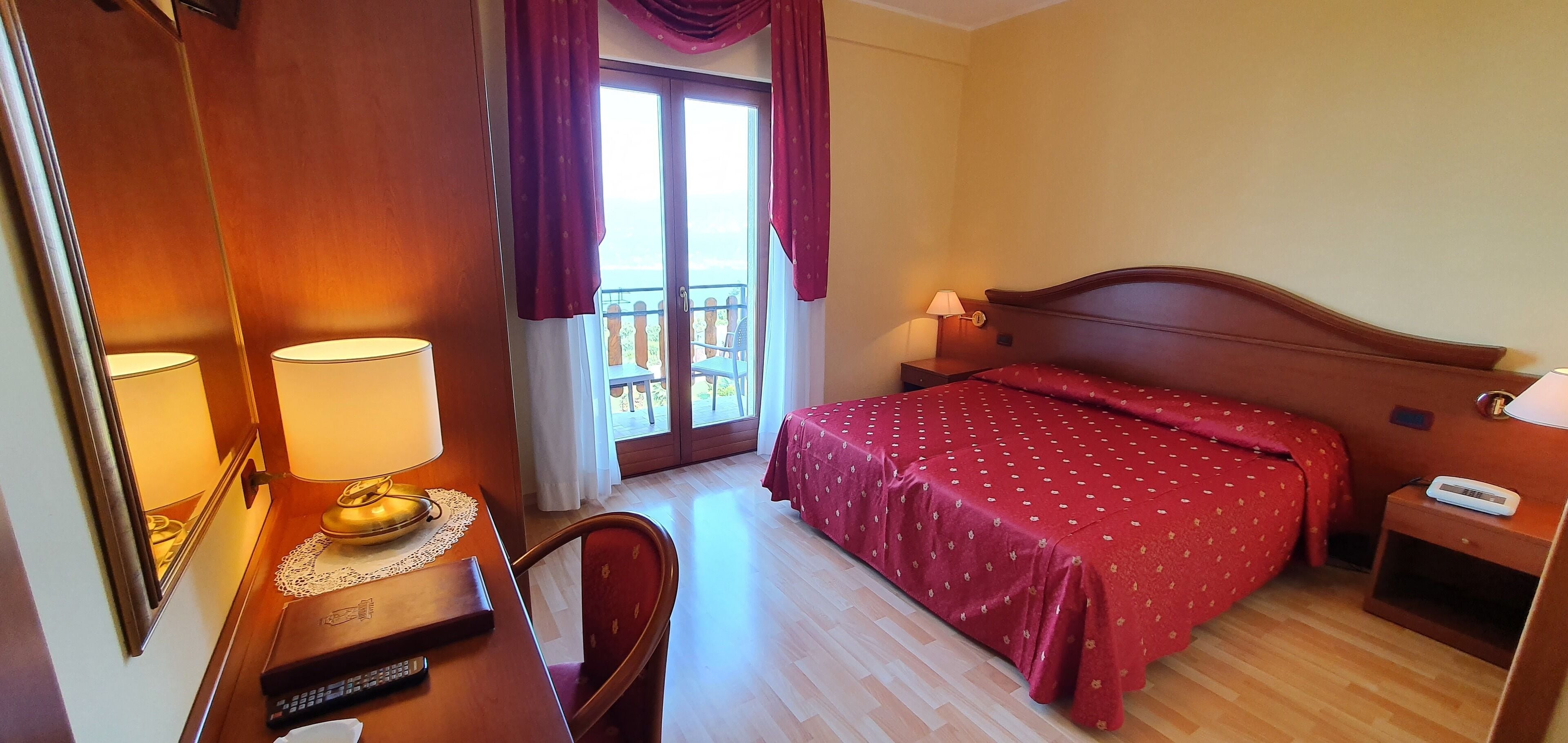 A hotel room with a wooden bed, a desk with a lamp, a chair, and a view of the outdoors through a glass door.