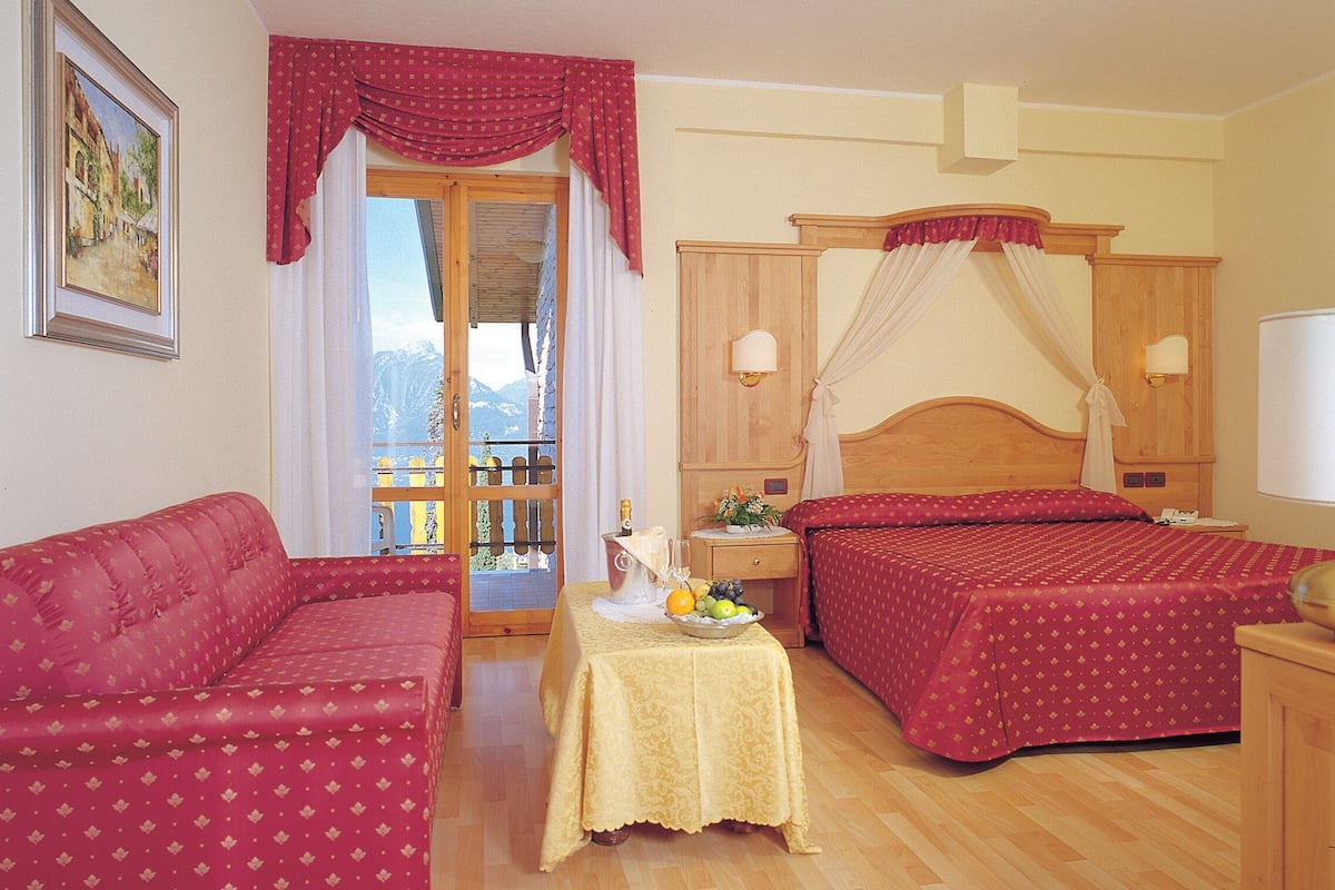 Habitación de hotel con dos camas, un sofá, una mesa con fruta y un balcón con vistas a la montaña.