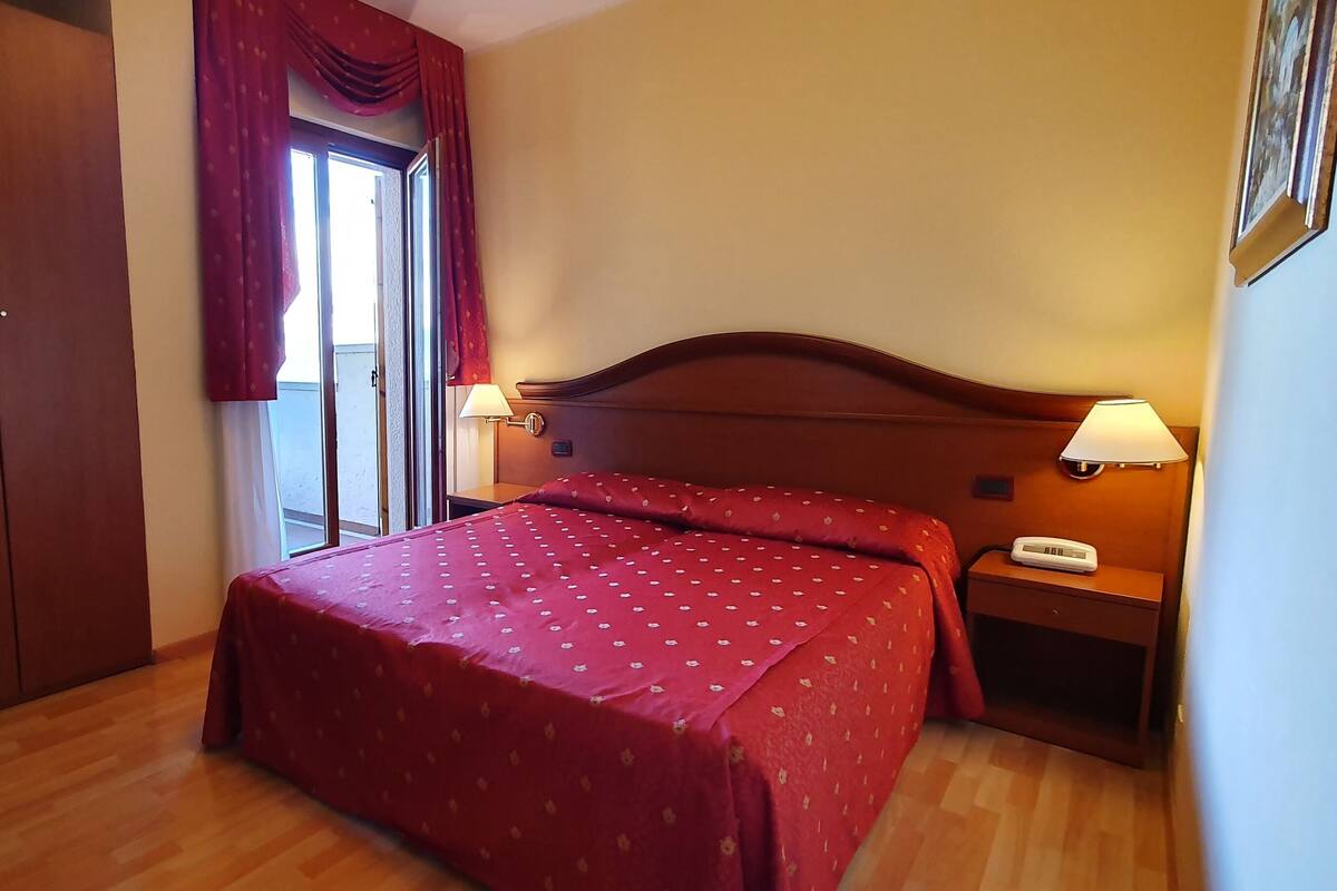 Una habitación de hotel con cama, mesita de noche, lámpara, armario y ventana con cortinas.