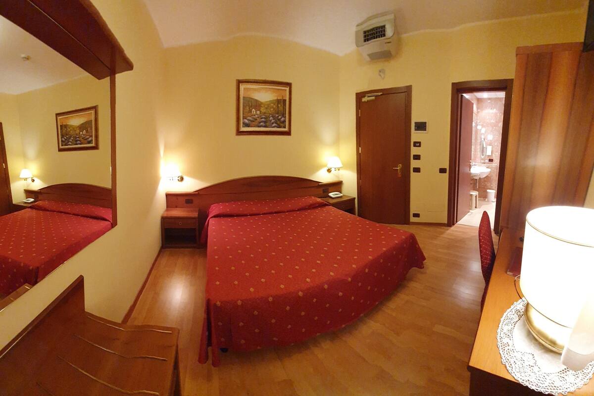 Habitación de hotel con dos camas individuales, suelo de madera, un escritorio con lámpara y un cuadro en la pared.