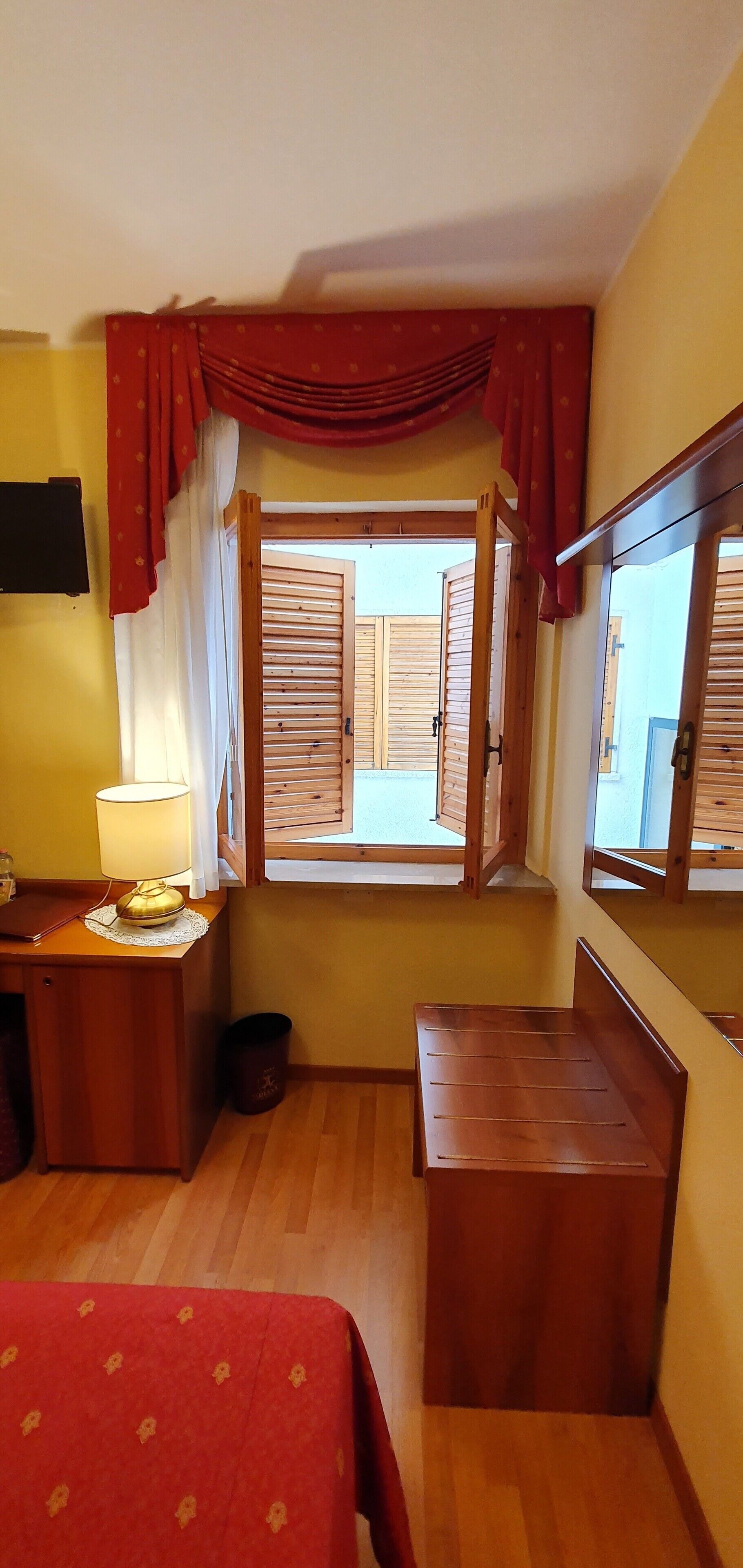 Un dormitorio con cama de madera, una mesita de noche con lámpara, una ventana con contraventanas de madera y una cortina roja.