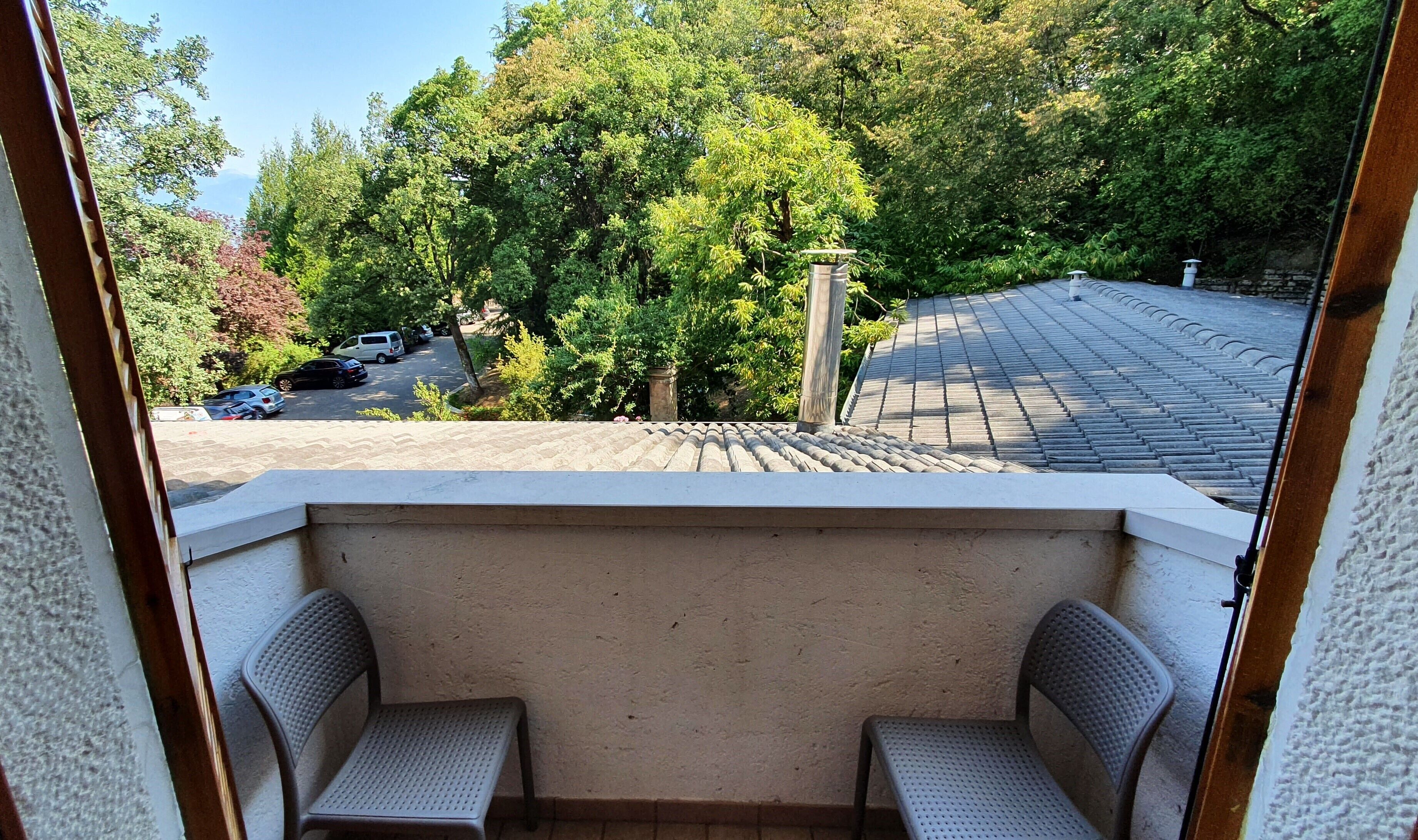 Un balcon avec deux chaises, une vue sur un parking avec des voitures et un jardin avec des arbres.