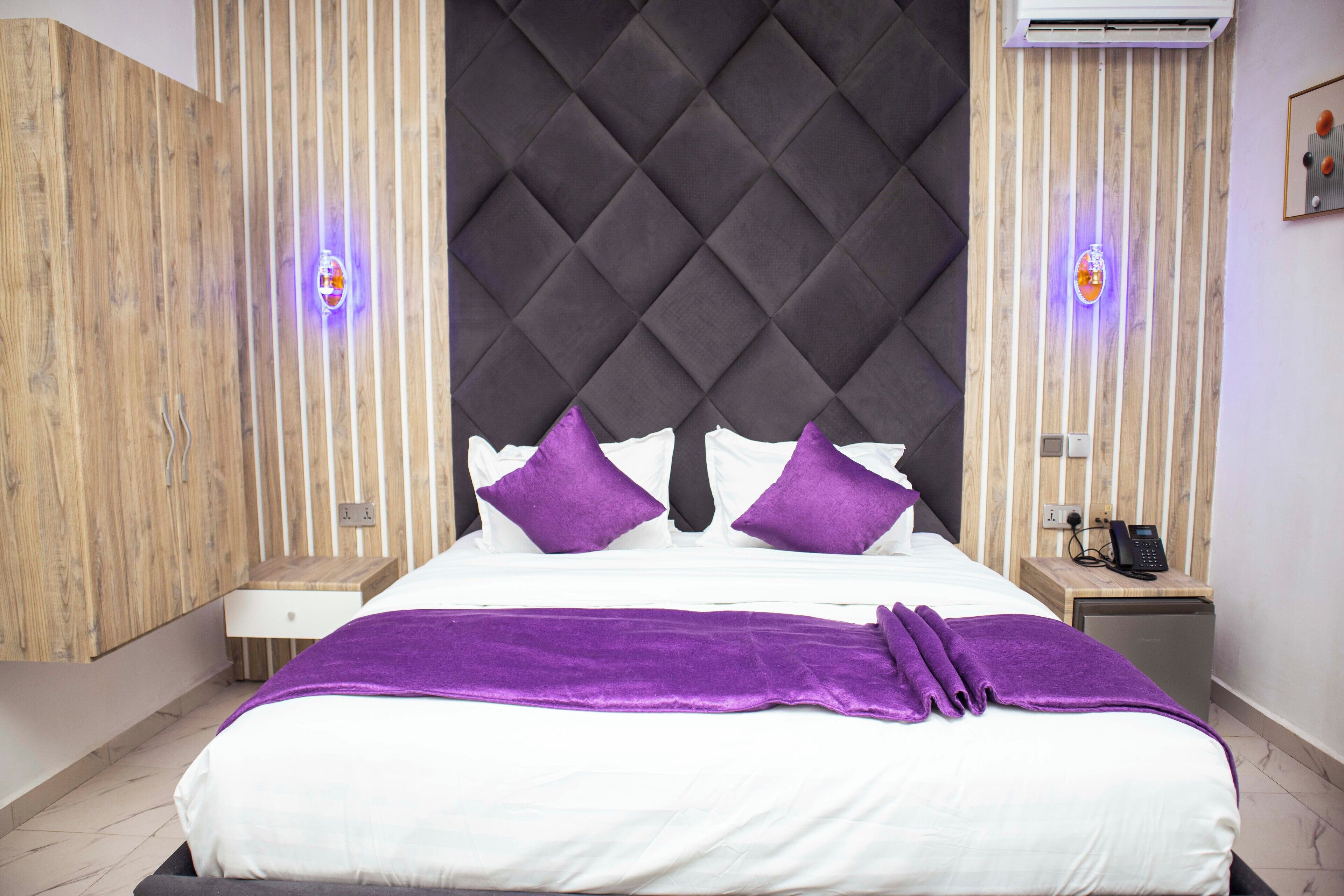 Una habitación de hotel moderna con una cama grande, un armario de madera, una mesita de noche pequeña y un buró con un teléfono.