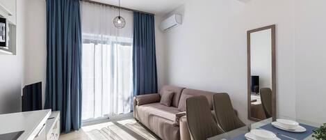 A modern hotel room with a sofa, a dining table with chairs, a large mirror, and a window with curtains.