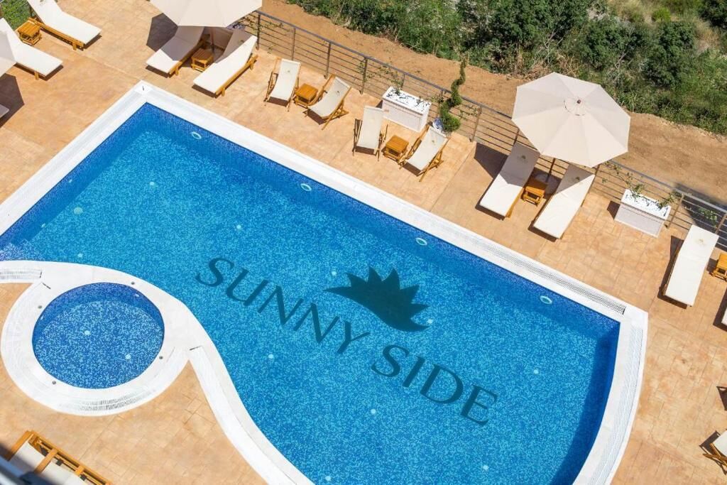A swimming pool with 'SUNNY SIDE' written on the water, surrounded by lounge chairs and umbrellas.