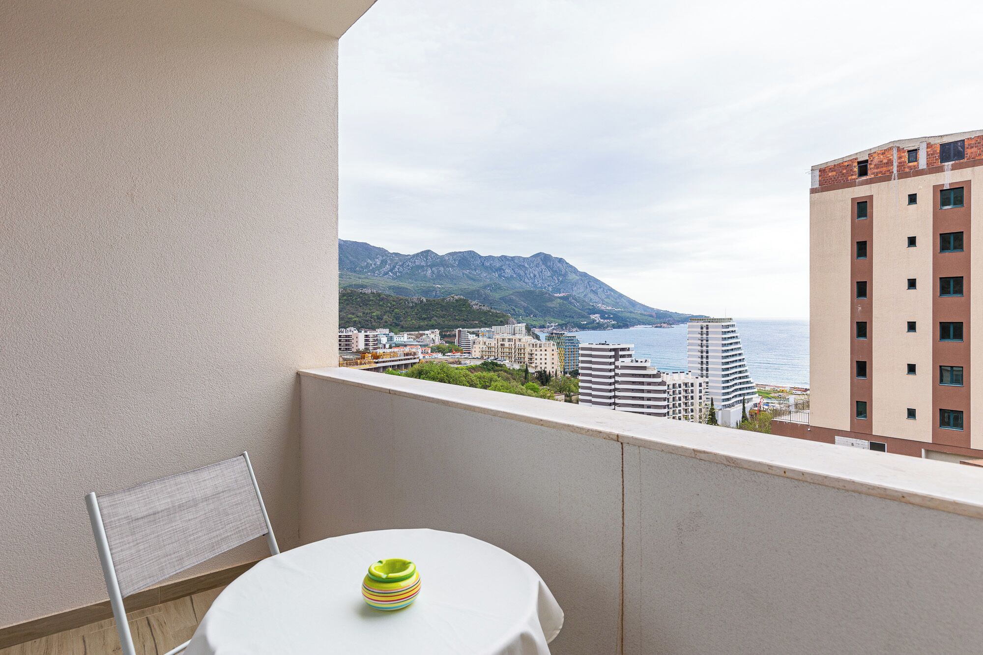 A balcony with a table and chair, overlooking a coastal cityscape with mountains in the distance.