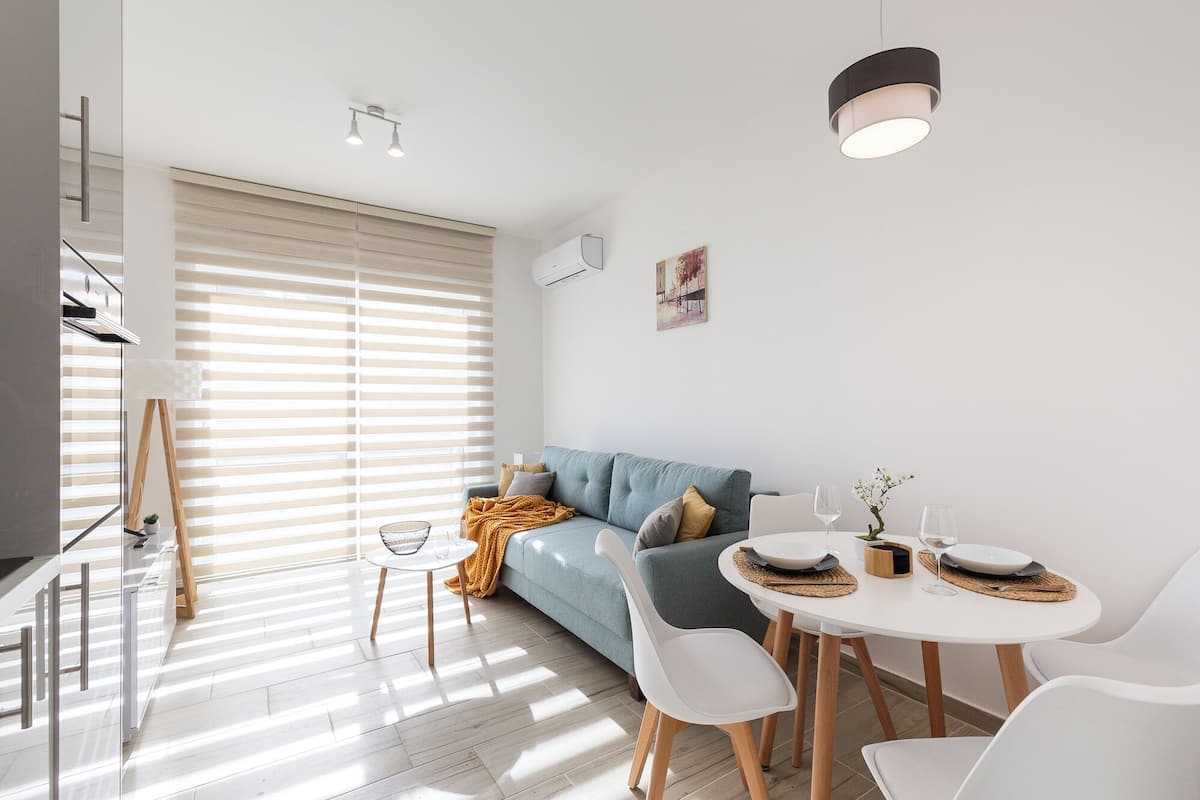 Ein modernes Wohnzimmer mit Sofa, Esstisch und Stühlen. An der Wand hängt ein Gemälde, ein Fenster mit Jalousien und eine Deckenleuchte.
