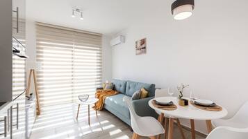 A modern living room with a sofa, dining table, and chairs. There is a painting on the wall, a window with blinds, and a ceiling light fixture.