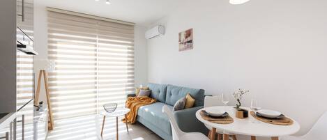 A modern living room with a sofa, dining table, and chairs. There is a painting on the wall, a window with blinds, and a ceiling light fixture.