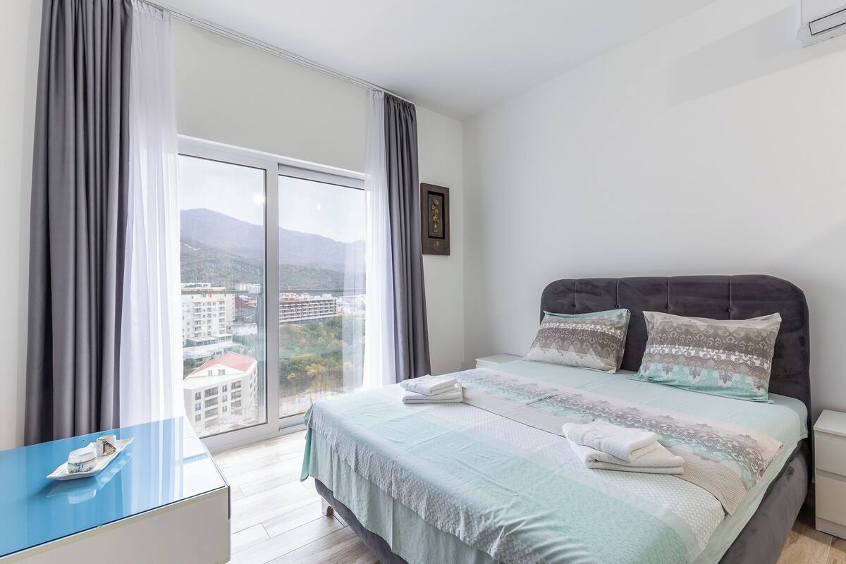 Ein Schlafzimmer mit einem Bett, einem Nachttisch, einem blauen Tisch und Blick auf Gebäude durch ein Fenster.
