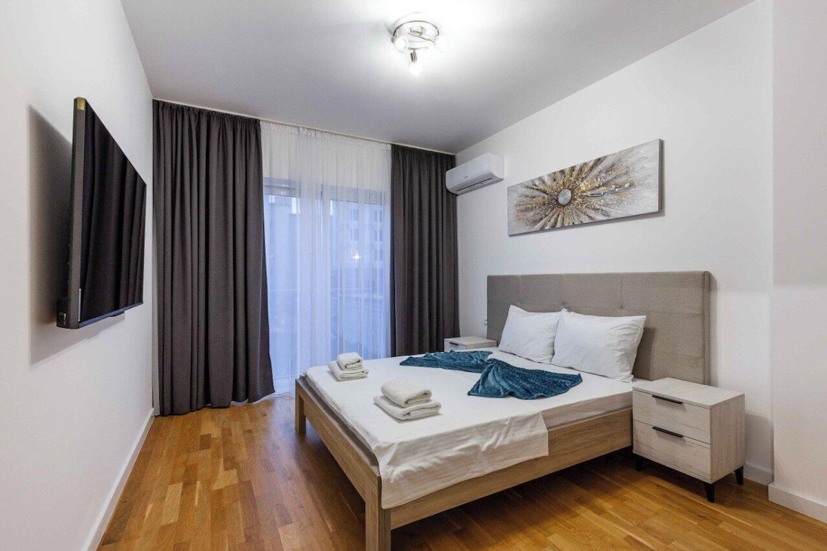Ein modernes Hotelzimmer mit einem großen Bett, einem Nachttisch, einem flachbildfernseher an der Wand und einem Fenster mit Vorhängen.