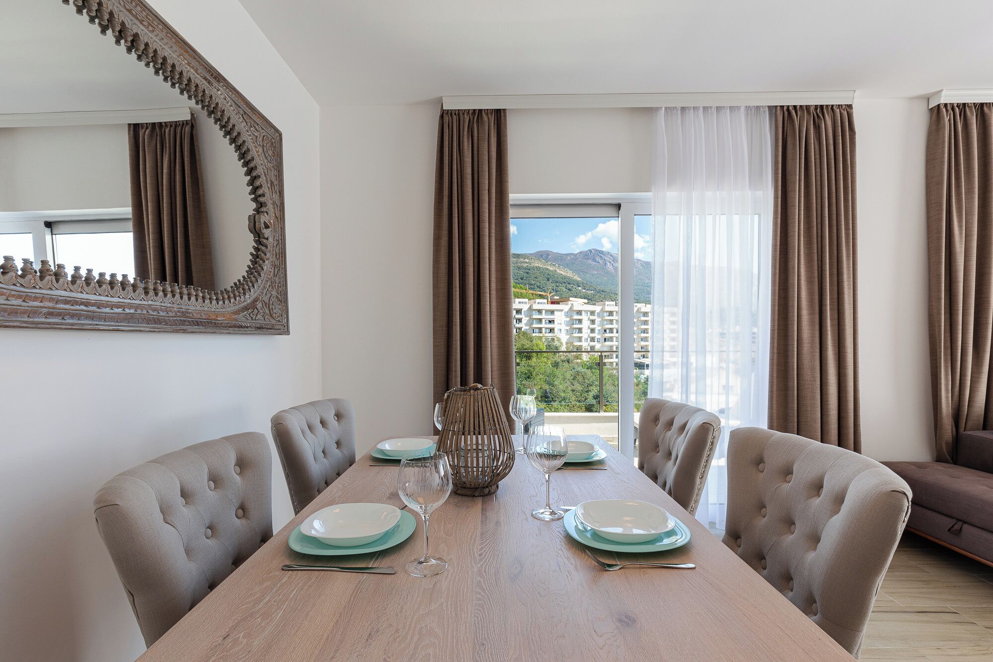 A dining room with a wooden table set for four, a large mirror, and a view of buildings and mountains through the window.