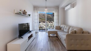 Un moderno salón con un televisor de pantalla plana, un sofá color beige y una pequeña mesa de centro redonda. Un amplio ventanal ofrece vistas a un paisaje montañoso y edificios de la ciudad.