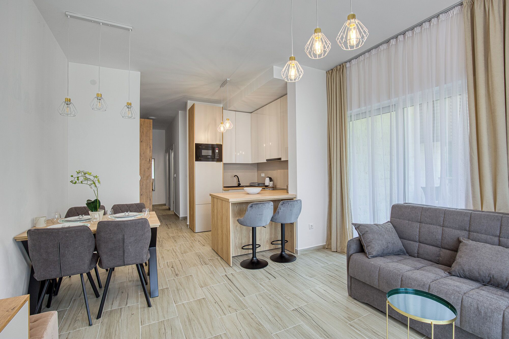 A modern living room with a grey sofa and a small round table, a dining area with a table and chairs, and a kitchen with a microwave and wooden cabinets.