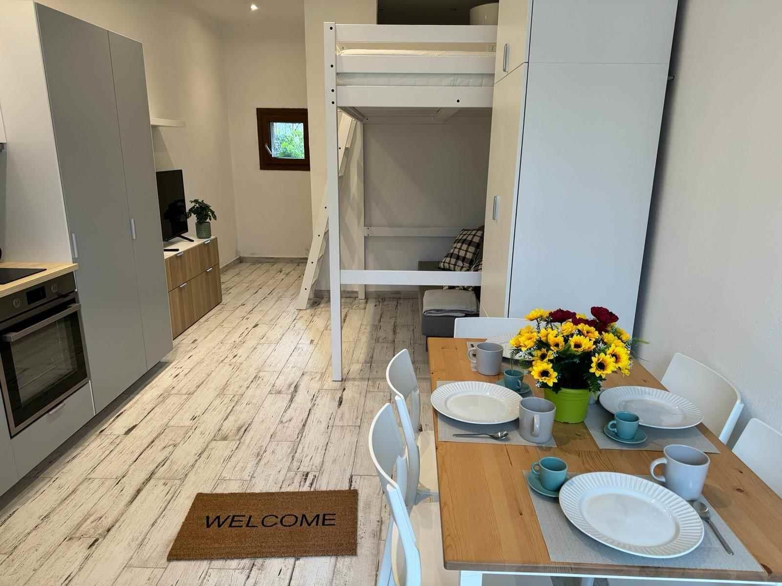 A compact living space with a bunk bed, kitchenette, dining area, and a welcome mat.