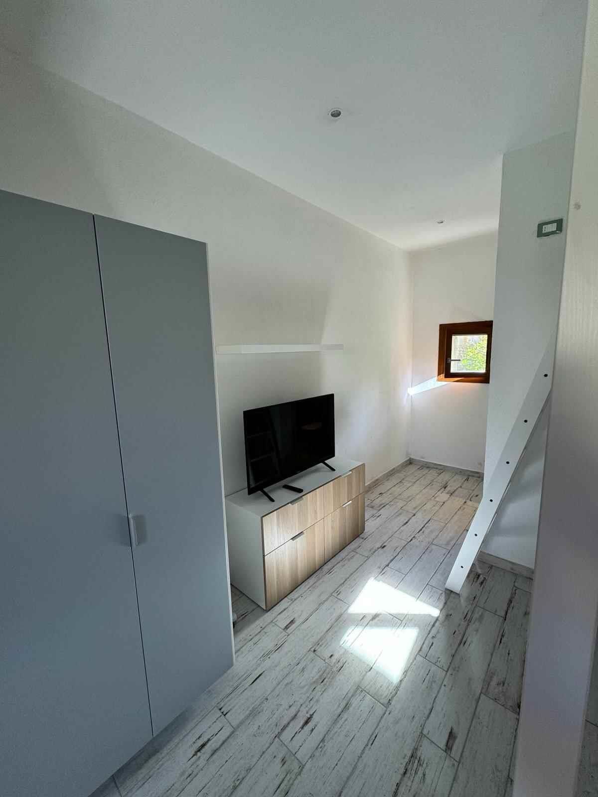 A modern, minimalistic room with a flat-screen TV on a wooden cabinet, a wardrobe, and a staircase.