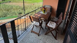 A balcony with a wooden table and chairs, a flower vase, and a view of a green landscape with houses.