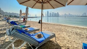 Una playa con sillas de descanso y sombrillas, un horizonte urbano al fondo y un mar tranquilo.