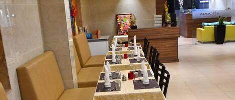 Un restaurant avec de longues tables disposées pour les repas, des banquettes sur les côtés et un plafond orné d’un ventilateur et de luminaires décoratifs.