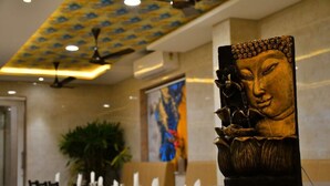A dining area with a Buddha statue centerpiece, a ceiling fan, and a large painting on the wall. - HOTEL ADHIKRISHNA ARCADE (Erode)