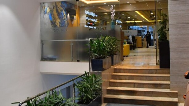 A modern hotel lobby with a staircase leading to the upper floor, potted plants, and a reception area with glass doors. - HOTEL ADHIKRISHNA ARCADE (Erode)
